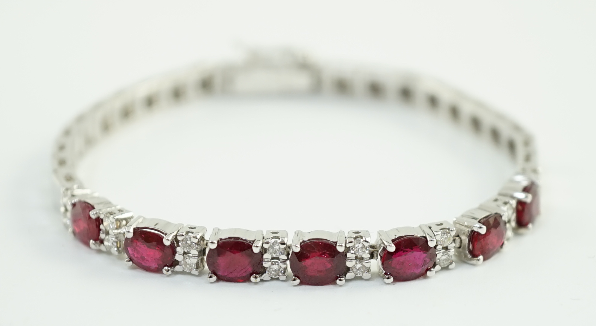 A 750 white gold and seven stone oval cut ruby set line bracelet, set with sixteen stone diamond spacers